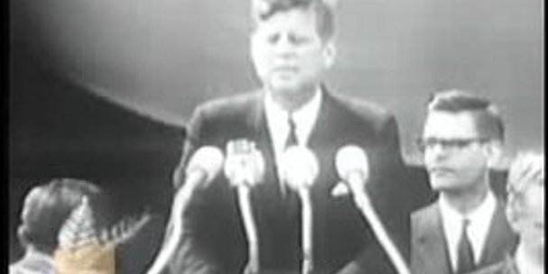 John F. Kennedy steht vor 4 Mikrofonen, im Hintergrund 3 Personen, schwarz weißes Bild