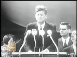 John F. Kennedy steht vor 4 Mikrofonen, im Hintergrund 3 Personen, schwarz weißes Bild