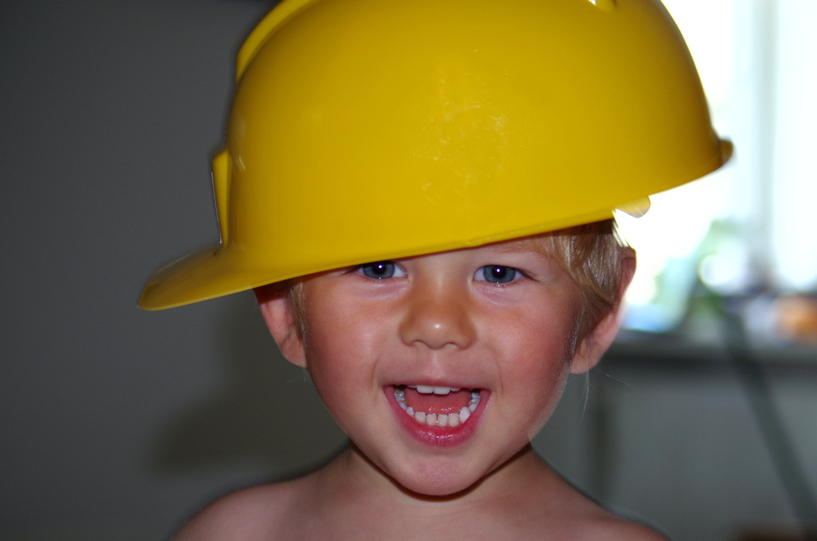 lächelndes Kleinkind, ca. 3 Jahre mit gelben Bauarbeiterhelm auf dem Kopf.